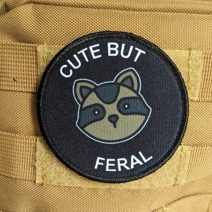 Peut inclure: Patch noir avec du texte blanc qui dit "Cute But Feral" et un raton laveur de dessin animé avec des yeux noirs et un nez brun.