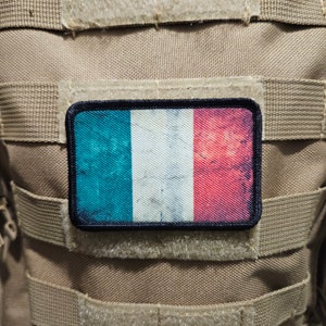 May include: A tan tactical vest with a velcro patch featuring the French flag. The flag is blue, white, and red with a distressed look.