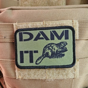 Pode incluir: Um remendo verde e preto com o texto "DAM IT" e uma silhueta preta de um castor.