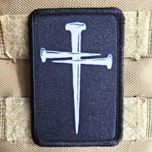 May include: A rectangular patch with a black background featuring a white nail cross design. The patch is bordered with black stitching and is attached to a tan fabric with hook and loop fasteners.