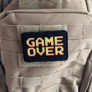May include: A black patch with white stitching and the words "GAME OVER" in yellow pixelated letters with red outlines. The patch is attached to a beige tactical backpack.