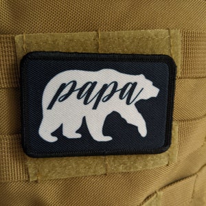 May include: A black and white embroidered patch featuring a white bear silhouette and the word "papa" in cursive. The patch is attached to a brown fabric background.