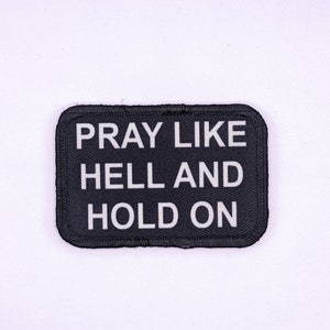 Puede incluir: Parche de tela negro con texto blanco que dice "Pray Like Hell And Hold On".