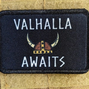 May include: Black rectangular patch with the words "VALHALLA AWAITS" in white. A Viking helmet graphic in tan, brown, and gold is centered between the words. The patch is on a tan fabric background.
