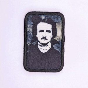 May include: Black and white embroidered patch featuring a portrait of Edgar Allan Poe. The patch has a black border and a blue and white background.