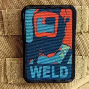 Può includere: Toppa rettangolare con un disegno di maschera da saldatore nei toni del blu, arancione e verde acqua. La parola "WELD" è stampata in blu in basso. La toppa ha un bordo nero ed è attaccata a un tessuto beige.