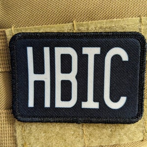 Op de afbeelding: Een rechthoekige patch met de witte letters "HBIC" op een zwarte achtergrond. De patch is bevestigd op een beige stof en heeft een klittenbandrug.