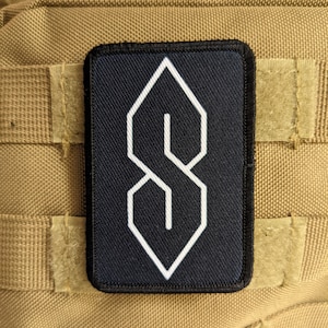 以下が含まれることがあります： ダイヤモンドの形をした白いスタイリッシュな文字「S」が書かれた黒いパッチ。パッチはタン色の布地に付けられています