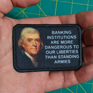 May include: Black fabric patch with a portrait of a man and the text "Banking institutions are more dangerous to our liberties than standing armies."