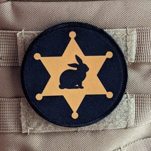 Pode incluir: Um remendo bordado preto e dourado com uma estrela de cinco pontas e uma silhueta de coelho dentro. O remendo é costurado em um tecido bege.