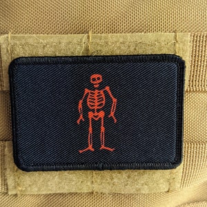 Pode incluir: Um remendo preto com um esqueleto vermelho bordado nele. O esqueleto está de pé com os braços ao lado do corpo. O remendo está preso a um fundo de tecido castanho.