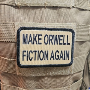 May include: A tan tactical vest with a black patch that reads "MAKE ORWELL FICTION AGAIN".