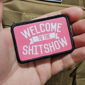 May include: A rectangular patch with the text "WELCOME TO THE SHITSHOW" in white on a pink background, framed by a black border. An American flag patch is visible in the background.