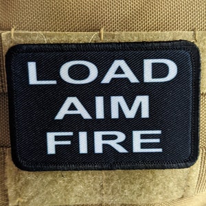 Può includere: Toppa nera con testo bianco che recita "LOAD AIM FIRE" su uno sfondo beige.