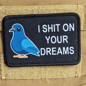 Puede incluir: Un parche negro con texto blanco que dice "I shit on your dreams". Un pájaro azul de dibujos animados está en el lado izquierdo del parche.