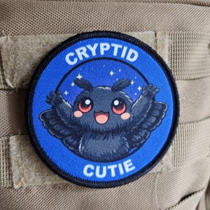 May include: A circular blue patch with the text "CRYPTID CUTIE" in white. The patch depicts a cartoon moth-like creature with a smiling face, outstretched arms, and star details. It is attached to a tan fabric.
