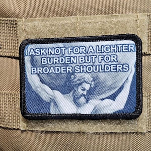 Könnte beinhalten: Ein schwarz-weißes Patch mit blauem Hintergrund, auf dem ein Mann ein großes Objekt hält. Der Text auf dem Patch lautet "I ask not for a lighter burden but for broader shoulders."
