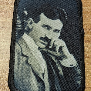 May include: Rectangular patch featuring a black and white portrait of Nikola Tesla. The image shows Tesla in a suit, with his hand near his face. The patch has a black border and is likely made of fabric.