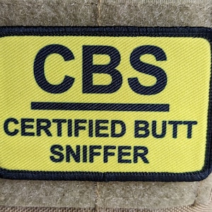 May include: A rectangular patch with a yellow background and black border. The patch features the letters "CBS" above the words "CERTIFIED BUTT SNIFFER" in black text. The patch is likely made of fabric.