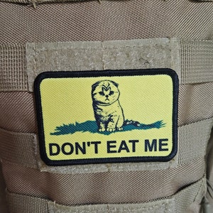 Puede incluir: Un parche amarillo con borde negro que presenta un gato de dibujos animados con el texto "DON'T EAT ME" en negro.