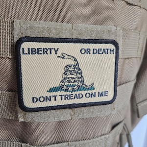 Puede incluir: Un chaleco táctico de color beige con un parche blanco y negro que dice "LIBERTY OR DEATH DON'T TREAD ON ME". El parche presenta una serpiente enroscada con una corona en la cabeza.