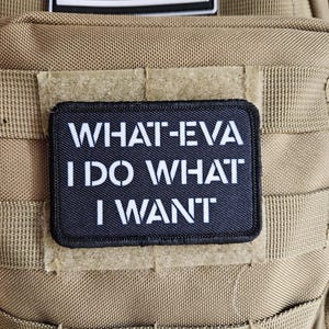 May include: A black rectangular patch with white text that reads "WHAT-EVA I DO WHAT I WANT" is displayed. Above it is a black and white American flag patch. The patches are attached to a tan tactical backpack.