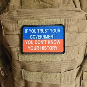Könnte beinhalten: Ein blauer und roter Patch mit dem Text "If you trust your government you don't know your history" auf einem braunen taktischen Rucksack.