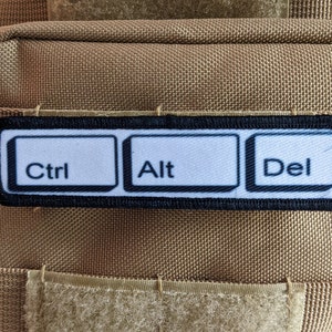 Könnte beinhalten: Ein schwarzer Patch mit weißem Text, der "Ctrl", "Alt" und "Del" in einer Reihe zeigt, ähnlich wie Computer-Tastaturtasten.