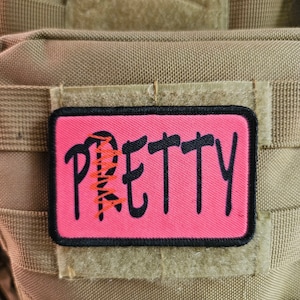 Puede incluir: Un parche rosa con costuras negras y la palabra "PRETTY" escrita en letras cursivas negras. El parche está unido a un chaleco táctico marrón.
