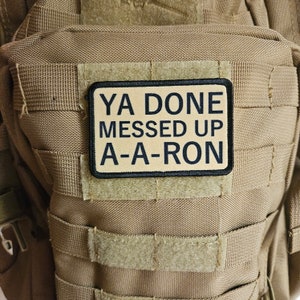 May include: A tan tactical backpack with a black and tan patch that reads "YA DONE MESSED UP A-A-RON".