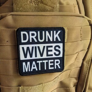 Puede incluir: Un parche blanco y negro con el texto "DRUNK WIVES MATTER" en una mochila táctica de color marrón.