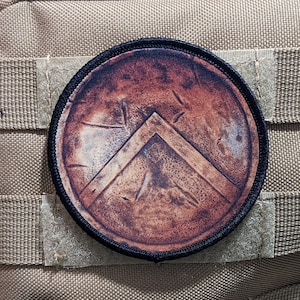 May include: A round, brown, and black patch with a stylized image of a shield with a chevron design. The shield has a distressed, weathered look.