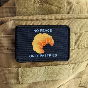 May include: A black patch with white text that reads "NO PEACE ONLY PASTRIES". A yellow croissant is in the center of the patch.