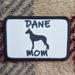 Może przedstawiać: Biała łatka z czarną obwódką, przedstawiająca czarną sylwetkę doga niemieckiego i tekst "DANE MOM".