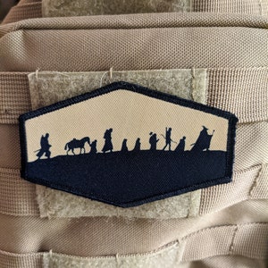 May include: A beige and black embroidered patch featuring a silhouette of a group of people walking with a horse. The patch is attached to a beige tactical vest.