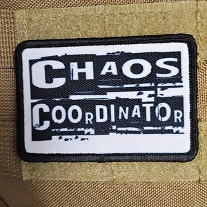 May include: Black and white patch with the text "CHAOS COORDINATOR" in a distressed font style. The patch is sewn onto a brown fabric background.