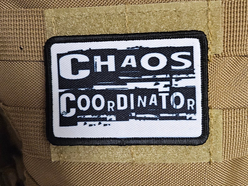 Peut inclure: Patch noir et blanc avec le texte "CHAOS COORDINATOR" dans un style de police distress&eacute;. Le patch est cousu sur un fond de tissu brun.
