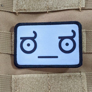 Puede incluir: Un parche blanco con un contorno negro y una cara de dibujos animados simple con las cejas levantadas y una boca en línea recta. La cara es un meme de Internet popular conocido como "feels bad man".