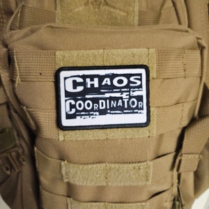 Peut inclure: Un &eacute;cusson blanc avec du texte noir qui dit "CHAOS COORDINATOR" sur un sac &agrave; dos tactique marron.