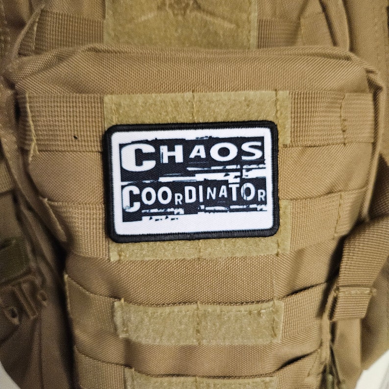 Peut inclure: Un &eacute;cusson blanc avec du texte noir qui dit "CHAOS COORDINATOR" sur un sac &agrave; dos tactique marron.