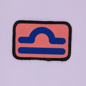 May include: A pink fabric patch with a blue Libra zodiac symbol. The patch has a black border.