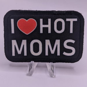 May include: Black rectangular patch with the text "I ❤️ HOT MOMS" in white letters. A red heart replaces the word "love". The patch has a black border and is displayed on a clear plastic stand.
