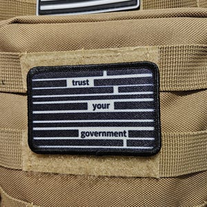 May include: A black and white patch with the words "trust your government" in a stylized font. The patch is attached to a tan tactical bag, with a black and white American flag patch above it.