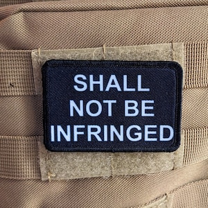 Könnte beinhalten: Ein schwarzer Patch mit weißem Text, der "SHALL NOT BE INFRINGED" lautet, ist auf einem braunen Stoffhintergrund befestigt.