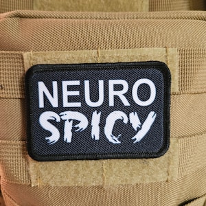 May include: A black patch with white text that reads "NEURO SPICY". The patch is sewn onto a tan fabric background.