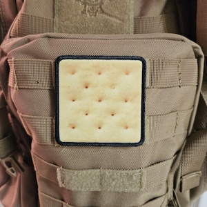 Może przedstawiać: Taktczny plecak w kolorze beżowym z czarną naszywką przedstawiającą brązowy krakers z wzorem kratki.