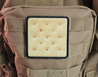 May include: A tan tactical backpack with a black patch featuring a brown cracker with a grid pattern.