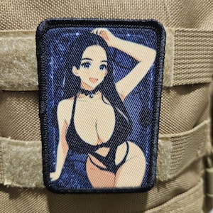Puede incluir: Un parche rectangular con una ilustración de estilo anime de una mujer en traje de baño negro. La mujer tiene el pelo largo y negro y posa con un brazo levantado. El fondo es azul oscuro con motas blancas y el parche tiene un borde negro.