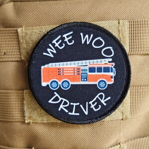 May include: A black and white round patch with the text "WEE WOO DRIVER" in white. A red and orange fire truck is in the center of the patch.