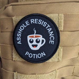 Może przedstawiać: Czarno-biała łatka z tekstem "ASSHOLE RESISTANCE POTION" w kółku wokół kreskówkowej filiżanki kawy z uśmiechniętą buzią i parą unoszącą się z niej.